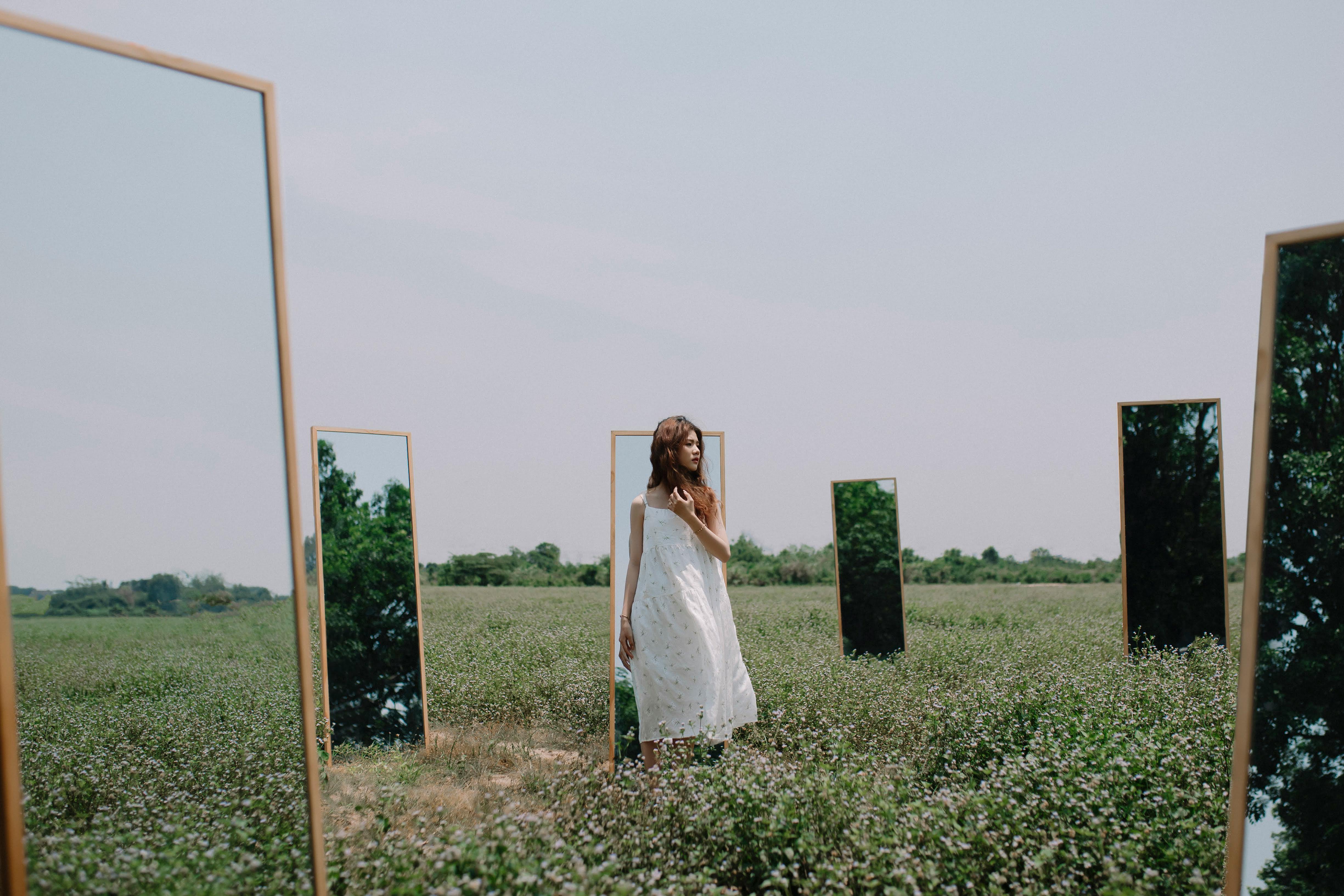 a photo of a Southeast Asian woman standing in a grass field surrounded by six haphazardly placed dressing mirrors